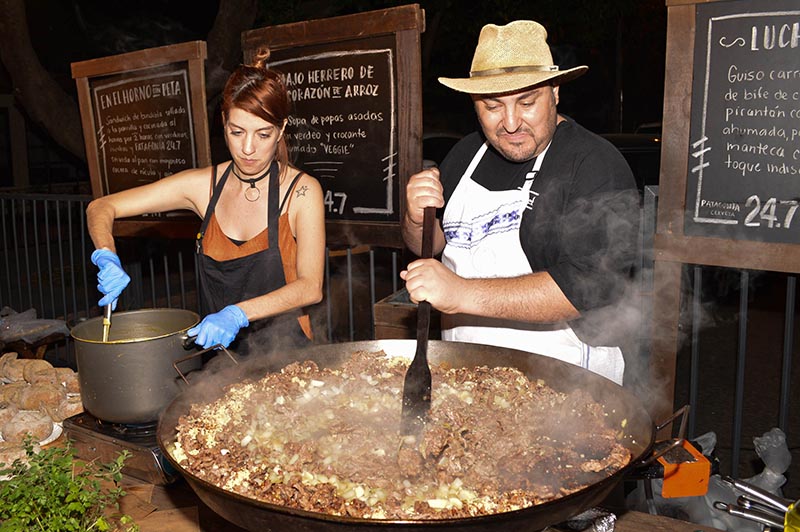 Luchef revolviendo con la pala lo que terminará siendo su famoso guiso carretero. A su lado, Majo Herrero preparando una sopa con papas asadas.