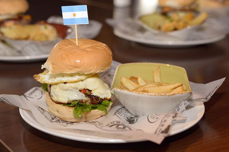 Tango Salsa Burger que representa a Buenos Aires. lleva rodajas de salchicha, salsa criolla, salsa alioli, queso Monterrey Jack, rúcula fresca y huevo frito.