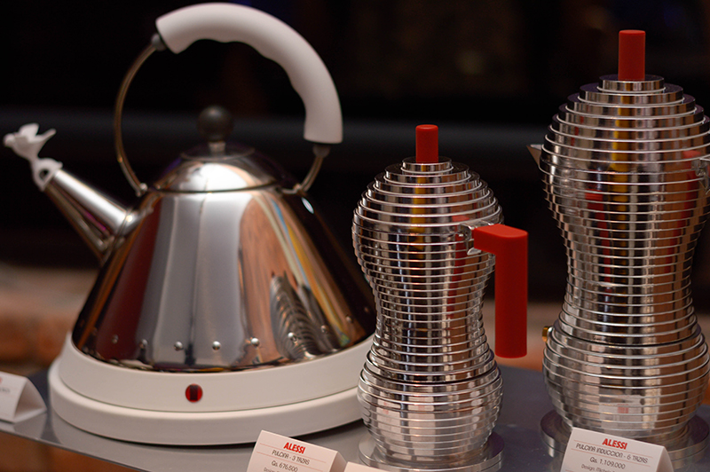 A la izquierda la cafetera Alessi, el producto más vendido de la marca. Tiene un mango colorido para indicar donde debe ir la mano y un pájaro en el pico. La forma cónica permite que pueda alcanzar altas temperaturas en poco tiempo.