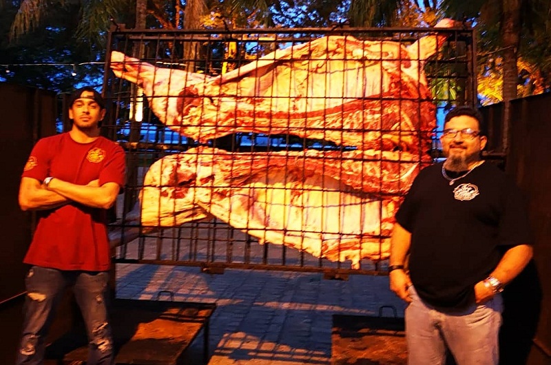 Leyzman Salim junto a un adyudante posa junto al novillo entero puesto a la parrilla para iniciar la cocción.