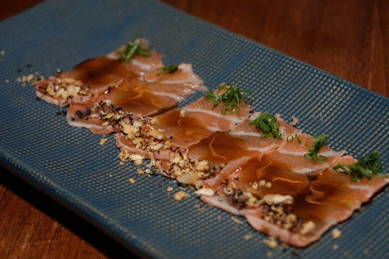 El tiradito OSK Tataki, finas láminas de salón sellado, con salsa cítrica oriental, togarashi, encurtidos de Kyuri y negi.