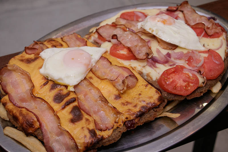 A la izquierda la milanesa Del Estudianhte (queso Cheddar, salsa barbacoa, panceta, huevo frito y papas fritas. A la derecha La Campeona El Antojo, muzzarella, jamon cocido, rodajas de tomate, cebolla, pancetas y papas fritas.